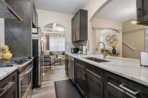 Una cocina moderna con electrodomésticos de acero inoxidable, encimera de mármol y vista hacia una sala de estar con sofá y mesa de centro.