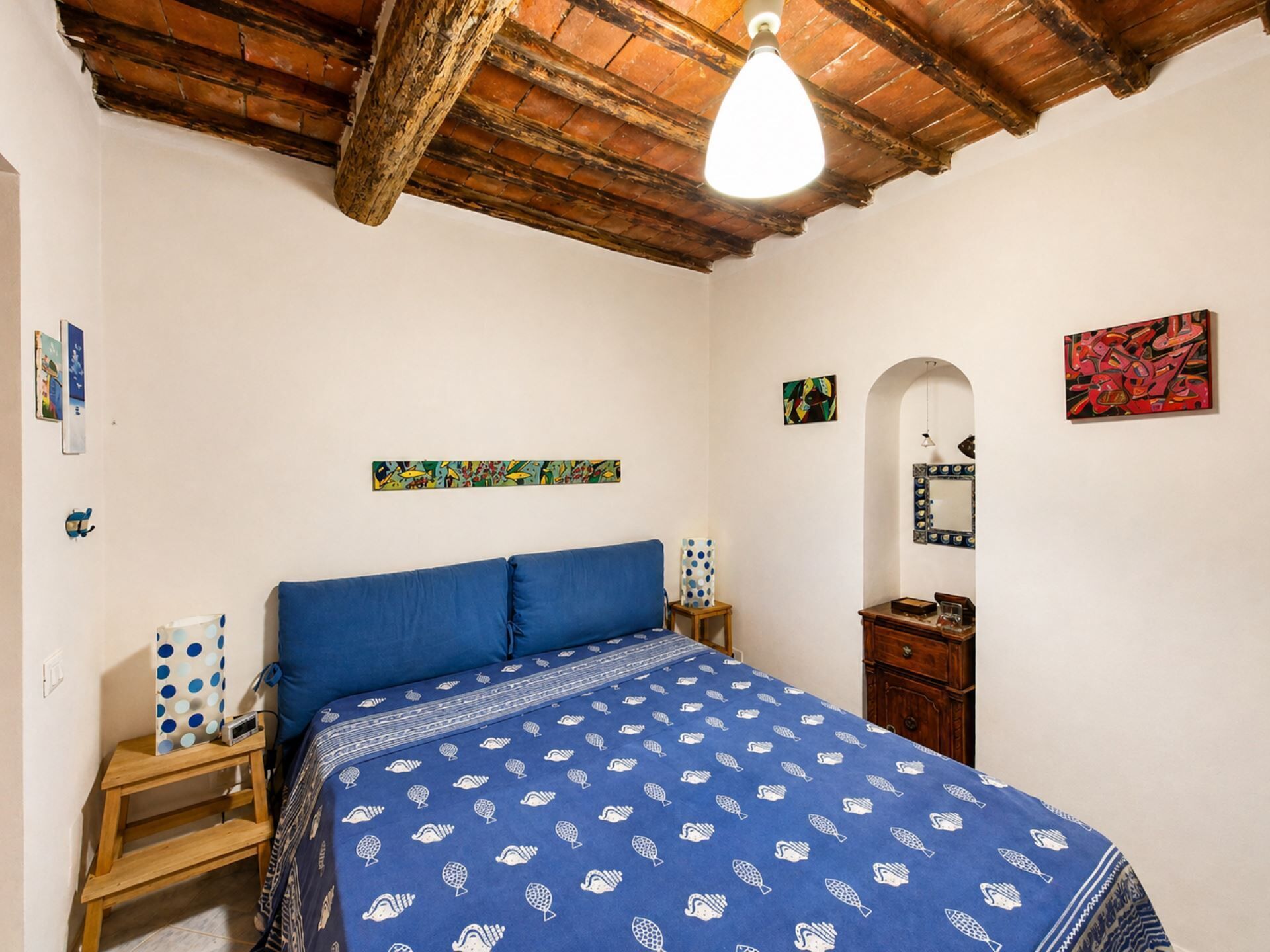 Un dormitorio con techo de madera, una cama con colcha azul estampada, una mesita de noche con lámpara de lunares y un aparador de madera.