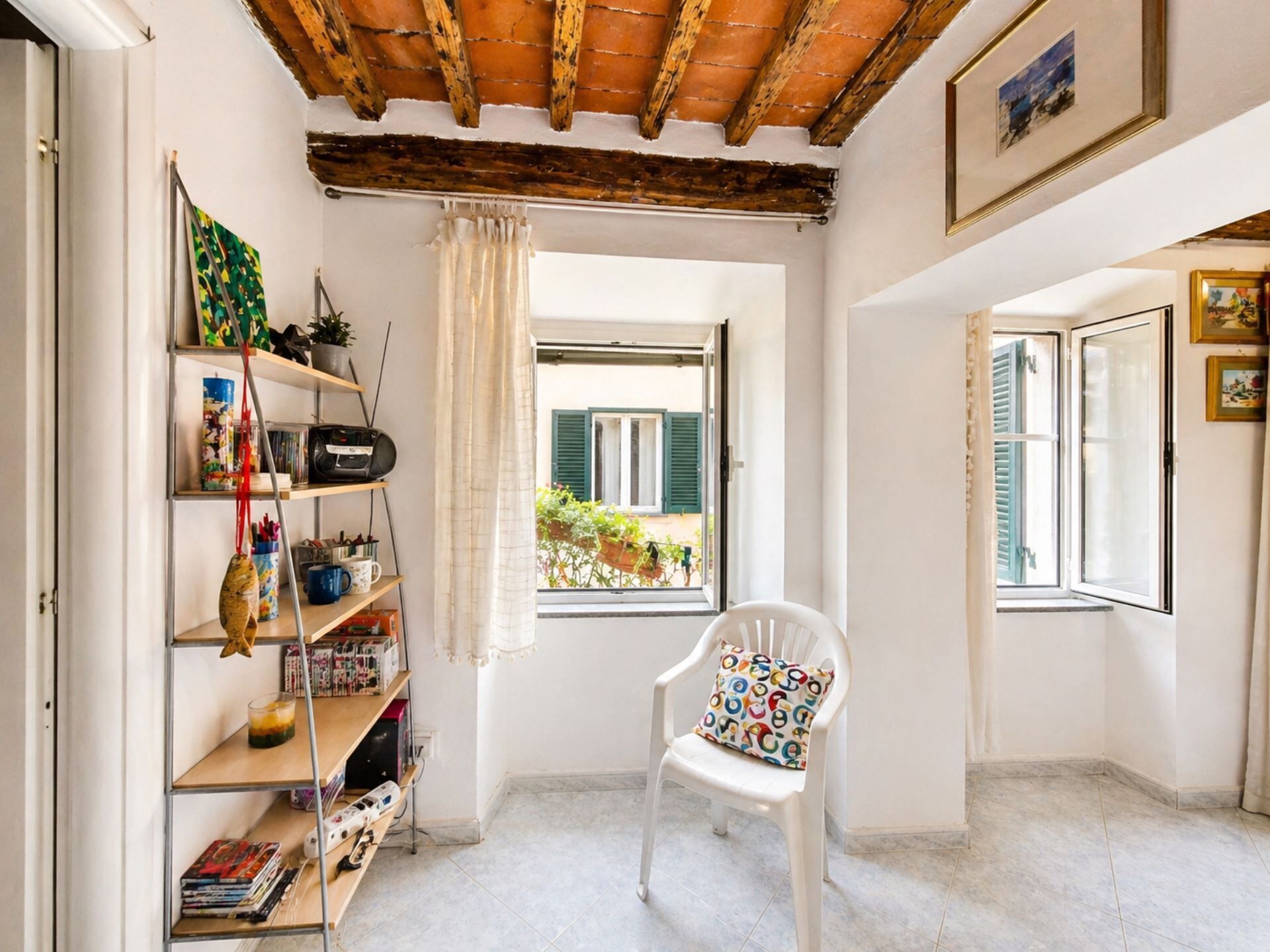 Une pièce avec un plafond en bois, une chaise blanche agrémentée d’un coussin coloré, une bibliothèque remplie de divers objets et une fenêtre donnant sur un paysage verdoyant.