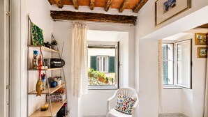Una habitación con techo de madera, una silla blanca con cojín de colores, una estantería con diversos objetos y una ventana que ofrece vistas al verdor exterior.