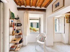 Ein Zimmer mit Holzboden, ein weißer Stuhl mit bunter Sitzkissen, ein Bücherregal mit diversen Gegenständen und ein Fenster mit Blick ins Grüne.