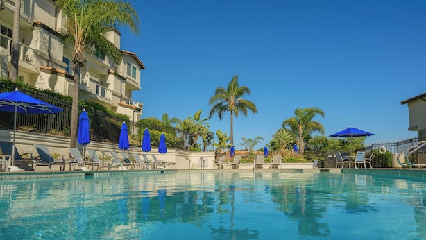 A swimming pool with lounge chairs and umbrellas, surrounded by palm trees and a building with balconies. - Sunny 2-bedroom condo in beautiful Laguna Niguel near best beaches in OC! (Laguna Niguel)