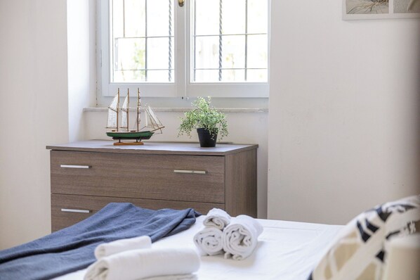 Un dormitorio con cama, cómoda, ventana con persianas, planta en maceta y toallas dobladas.