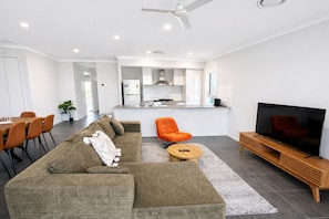 A modern living room with a sofa, a coffee table, an orange armchair, a TV, and a kitchen in the background. - Solara Retreat | Modern 4BR Home with Backyard (Warner)