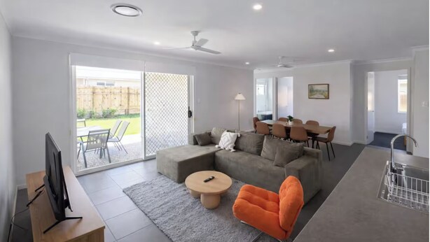En moderne stue med sofa, sofabord og fjernsyn, et spiseområde med stole, og et køkken med vask.