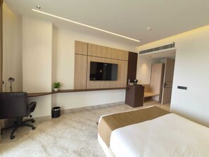 Quarto de hotel moderno com uma cama grande, uma escrivaninha, uma cadeira, uma TV fixada na parede e uma pequena planta.