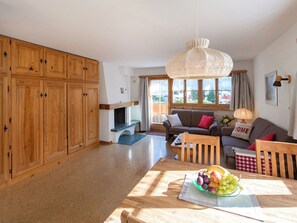 A spacious living area with wooden cabinets, a sofa, a dining table with fruit, and a large window. - Ferienhaus Muntschi, (Andeer), by Interhome (Andeer)