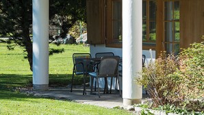 A covered outdoor seating area with a table and chairs, a wooden structure overhead, and a view of a grassy area with trees.