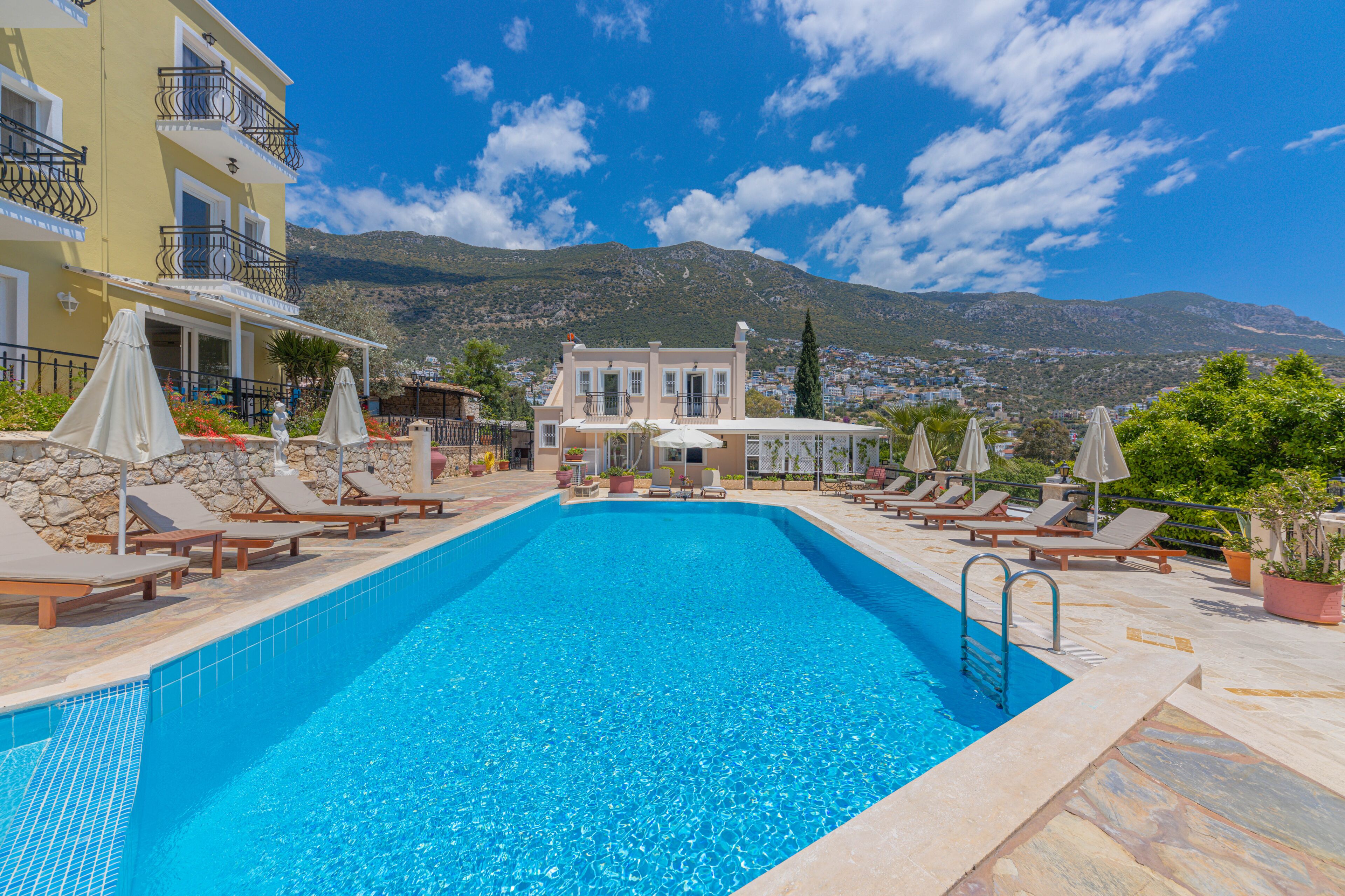 A swimming pool with lounge chairs and umbrellas, a building with balconies, and a mountainous backdrop.