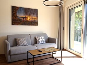A modern living room with a grey sofa, two patterned cushions, a wooden coffee table with a remote control, and a large window with a view of greenery. - Bergzeit Appartement Top 2 by Interhome (Saalbach-Hinterglemm)