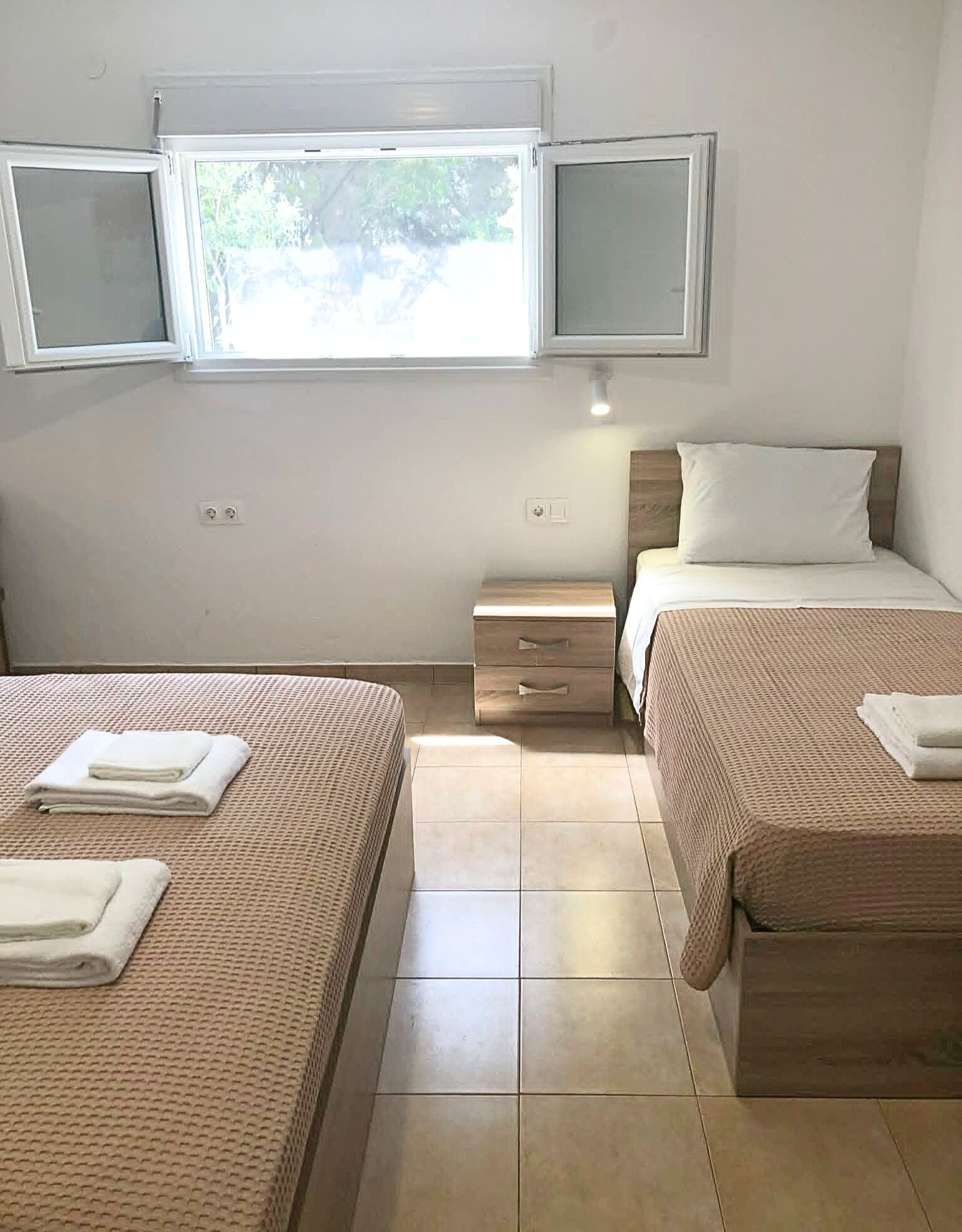 A hotel room with two beds, a small bedside table, a nightstand, a window with blinds, and a wall-mounted TV.
