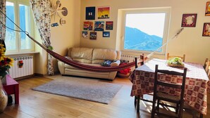 Una sala de estar acogedora con una hamaca, una mesa de comedor con mantel estampado y vistas a montañas a través de un ventanal grande.