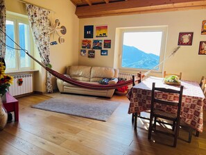 A cozy living room with a hammock, a dining table with a patterned tablecloth, and a view of mountains through a large window. - Farfa nature farmhouse with BBQ, Ping Pong and Fields for walkers (Castelnuovo di Farfa)
