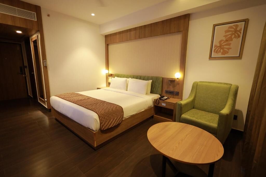 Una habitación de hotel con cama, mesita de noche, sillón verde y una mesa redonda de madera.