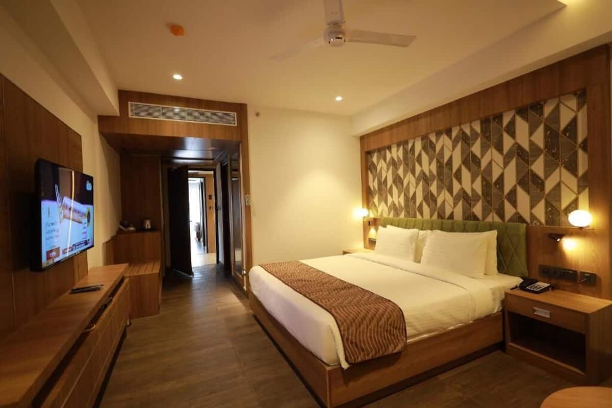 A hotel room with a bed, a television, a bedside table, a nightstand, and a wall-mounted light fixture.