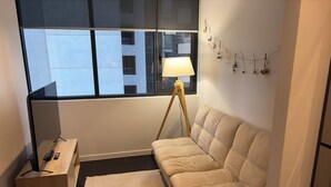 A room with a sofa, a lamp, a small table, a rug, and a window with a view of buildings. - Cozy Condo near Southern Cross / Pool & GYM (Melbourne)