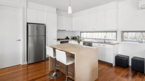 A modern kitchen with white cabinets, a stainless steel refrigerator, a wooden island with bar stools, and a window providing natural light. - Premium Location Family and Pet Friendly Property (Torquay)