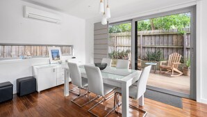 A modern dining area with a glass table, white chairs, and a view of a wooden deck and garden. - Premium Location Family and Pet Friendly Property (Torquay)