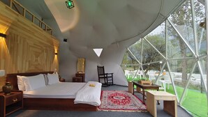 A spacious tent-style bedroom with a large bed, a wooden nightstand, a chair, a small table, and a view of the outdoors through large windows. - North Cliff by Stay Pattern (Gund)