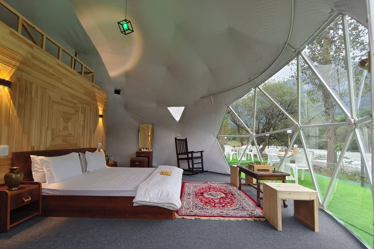 Um quarto em estilo tenda espaçoso, com uma cama grande, uma mesa de cabeceira de madeira, uma cadeira, uma mesinha e vista para o exterior através de amplas janelas.