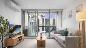 Sala moderna com sofá, mesa de centro e televisão, varanda com vista para edifícios e um vaso de planta.
