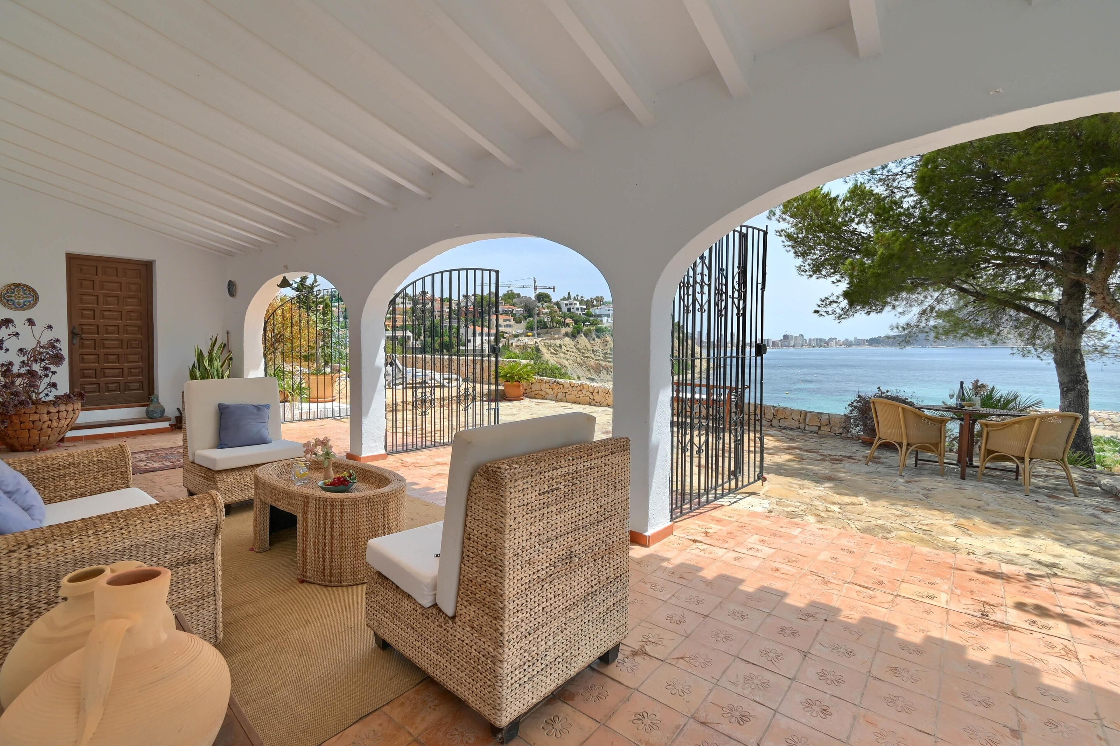 A sunlit patio with wicker furniture, a view of the sea, and a stone pathway leading to the beach.