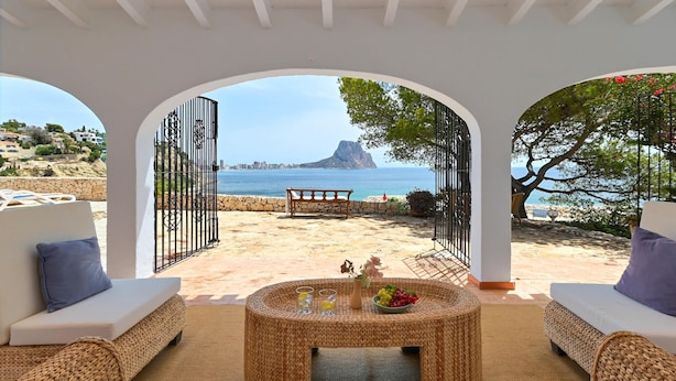 A beachside patio with wicker furniture, a round table with drinks and fruit, and a view of the sea and a mountain. - Casa Timeris 8 pax - Calpe, Costa Blanca (Calpe)
