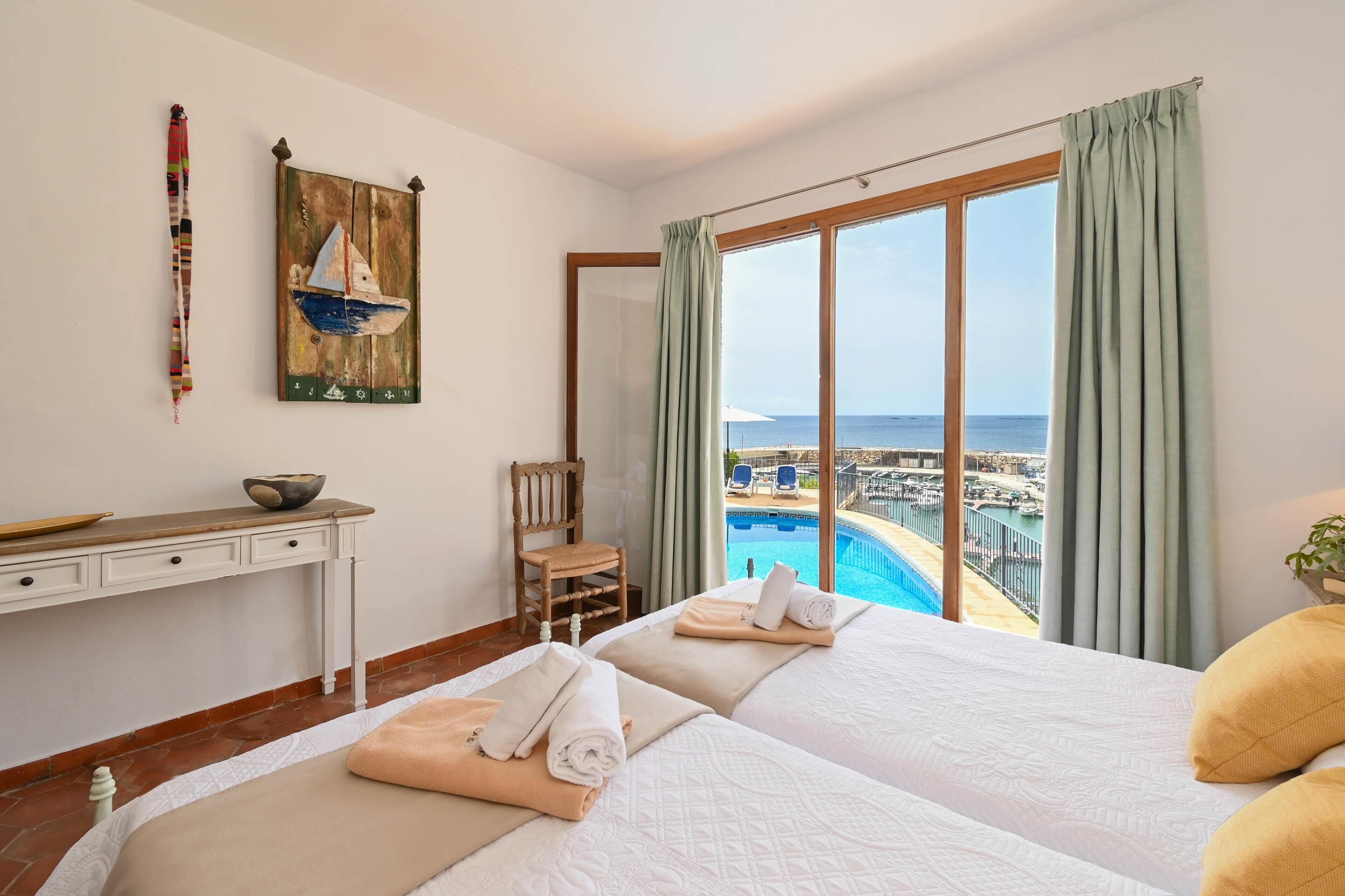 A hotel room with two beds, a desk, a chair, a painting, and a view of a marina and pool.