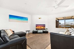 A living room with a flat-screen TV, a ceiling fan, a large window, and a beach-themed painting.