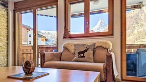 Un salon chaleureux avec une table en bois, un canapé offrant une vue sur les montagnes grâce à de grandes fenêtres, et un coussin décoratif.