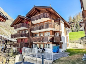 A multi-story building with wooden balconies, a covered outdoor seating area, and a grassy slope in the background. - Apartment Carlos by Interhome (Zermatt)