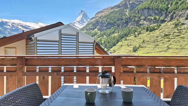Un balcon avec une table, des chaises et une cafetière, donnant sur un paysage montagneux avec un bâtiment en arrière-plan.