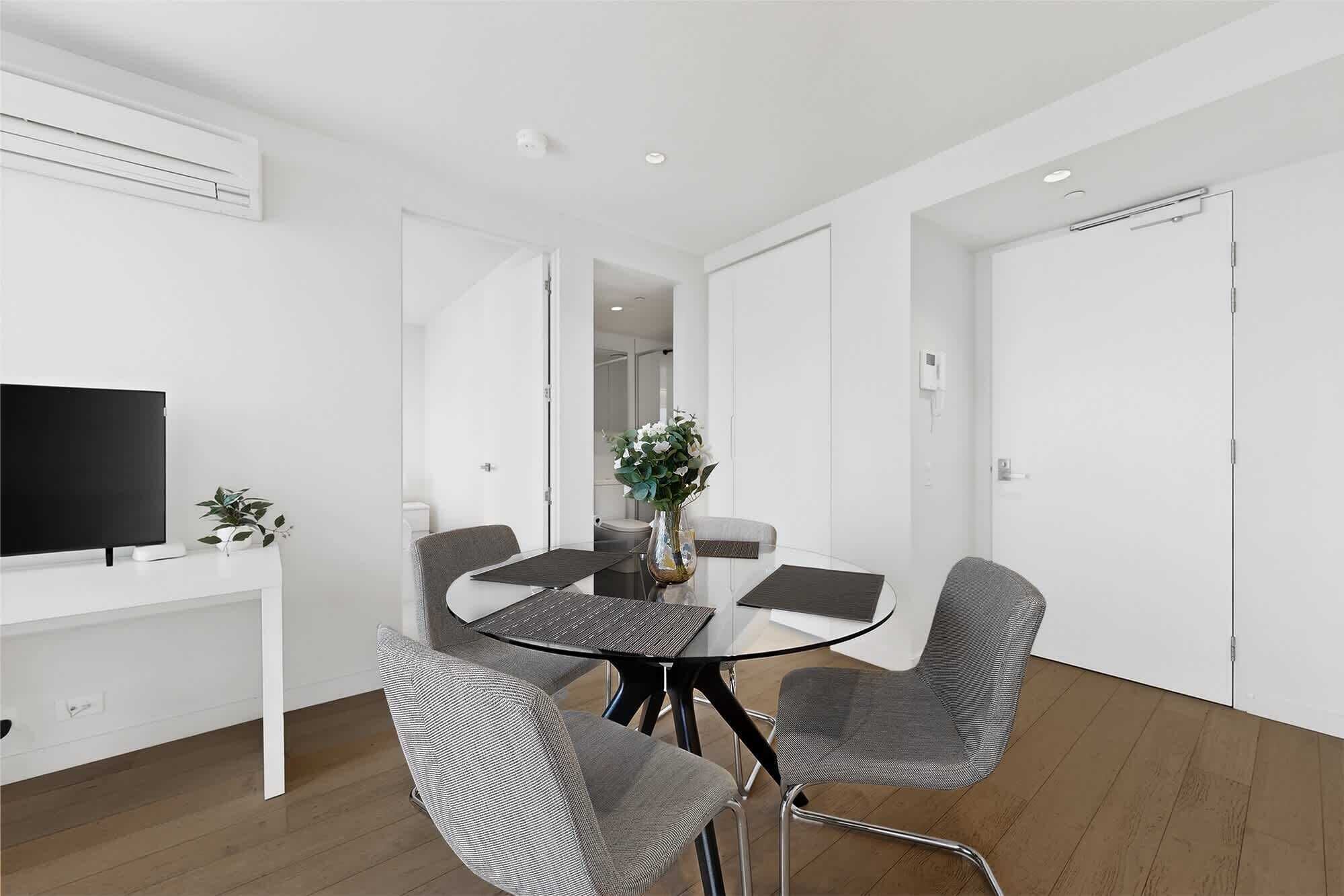 A modern room with a round table, four grey chairs, a flat-screen TV on a white stand, and a potted plant.