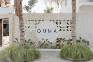 A hotel entrance with a white wall, palm trees, and a circular sign with 'QUMA Hotel & Restaurant' written on it. - Quma Boutique Hotel - Adults Only (Gili Trawangan)