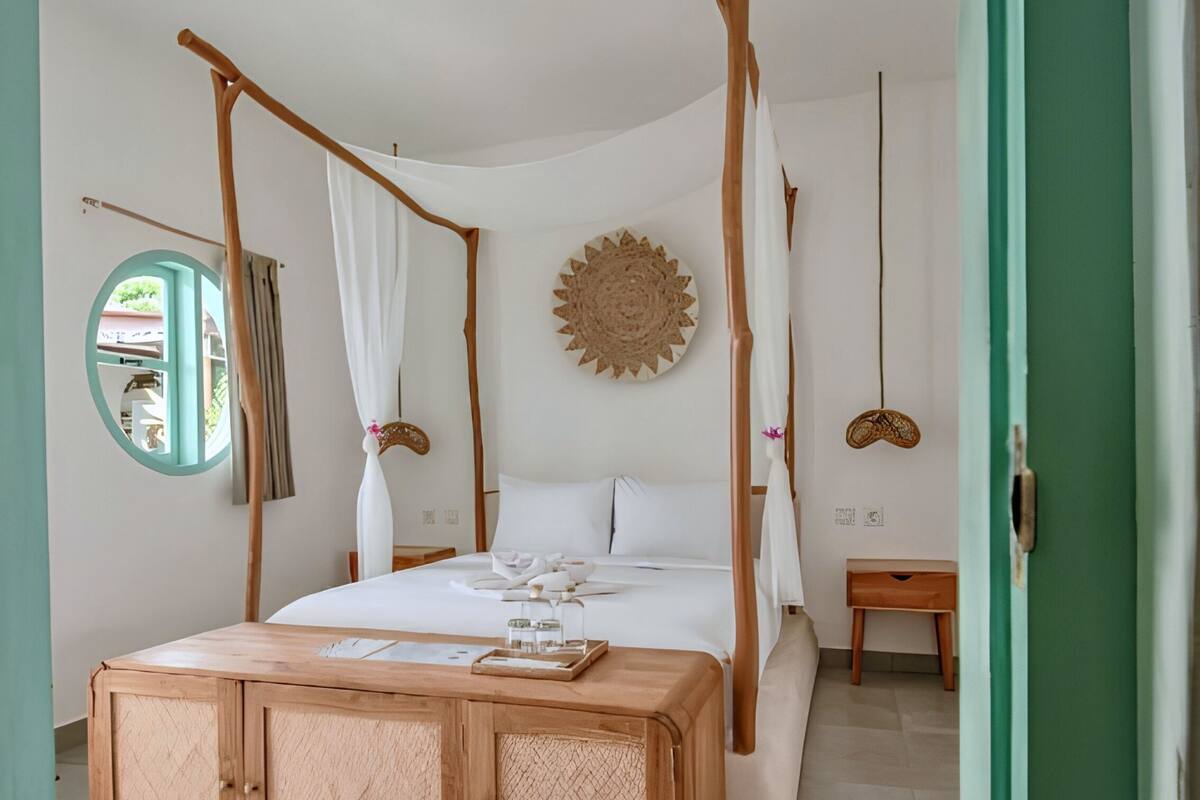 A four-poster bed with a canopy, a bedside table, a wall-mounted decorative piece, and a view of the outdoors through a window.