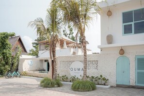 A modern building with a sign reading 'QUMA Hotel & Restaurant,' a thatched-roof structure, and a white wall with a circular logo. - Quma Boutique Hotel - Adults Only (Gili Trawangan)