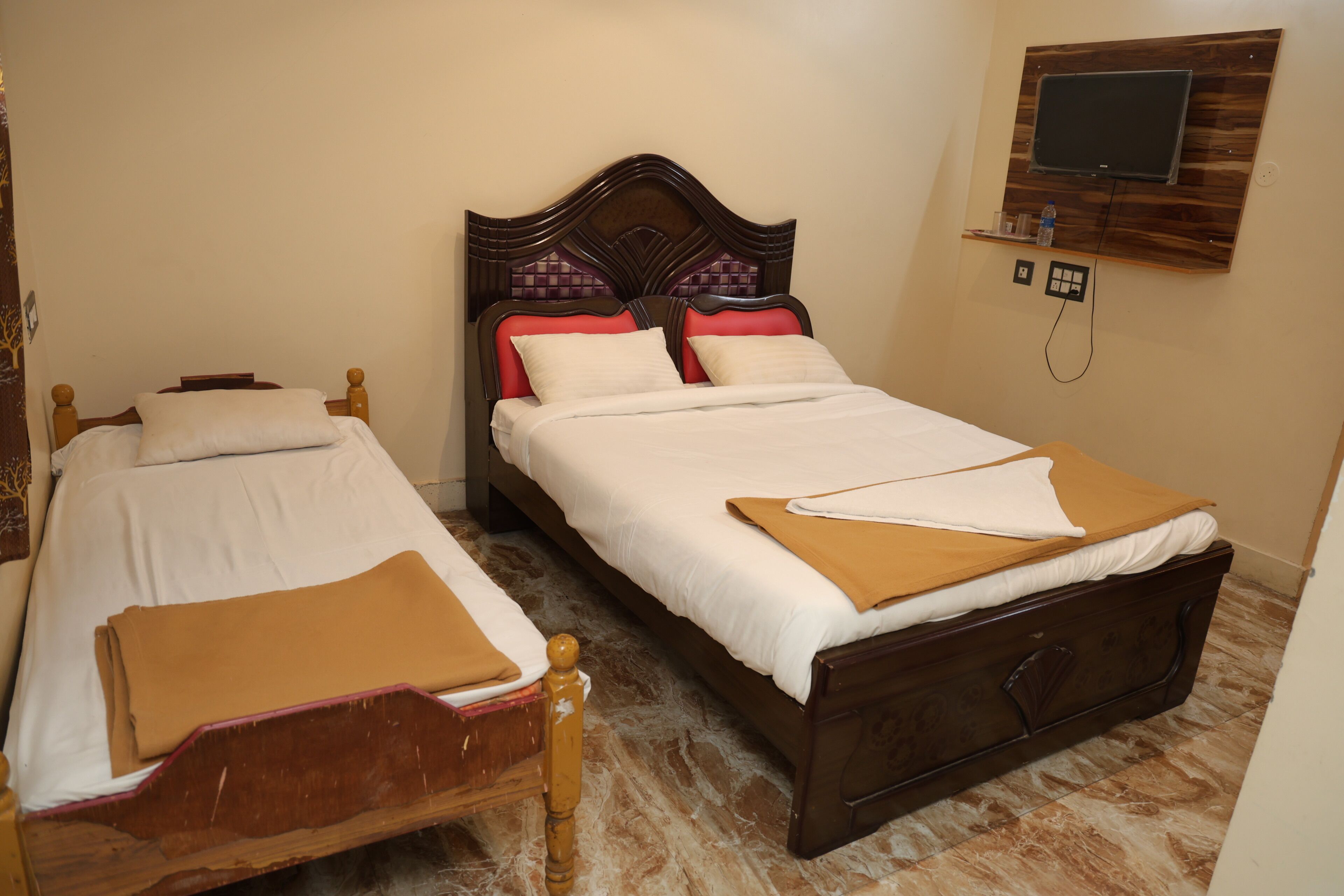 Two wooden beds with white linens and red pillows, a wooden headboard, a mounted TV, and a power outlet on the wall.
