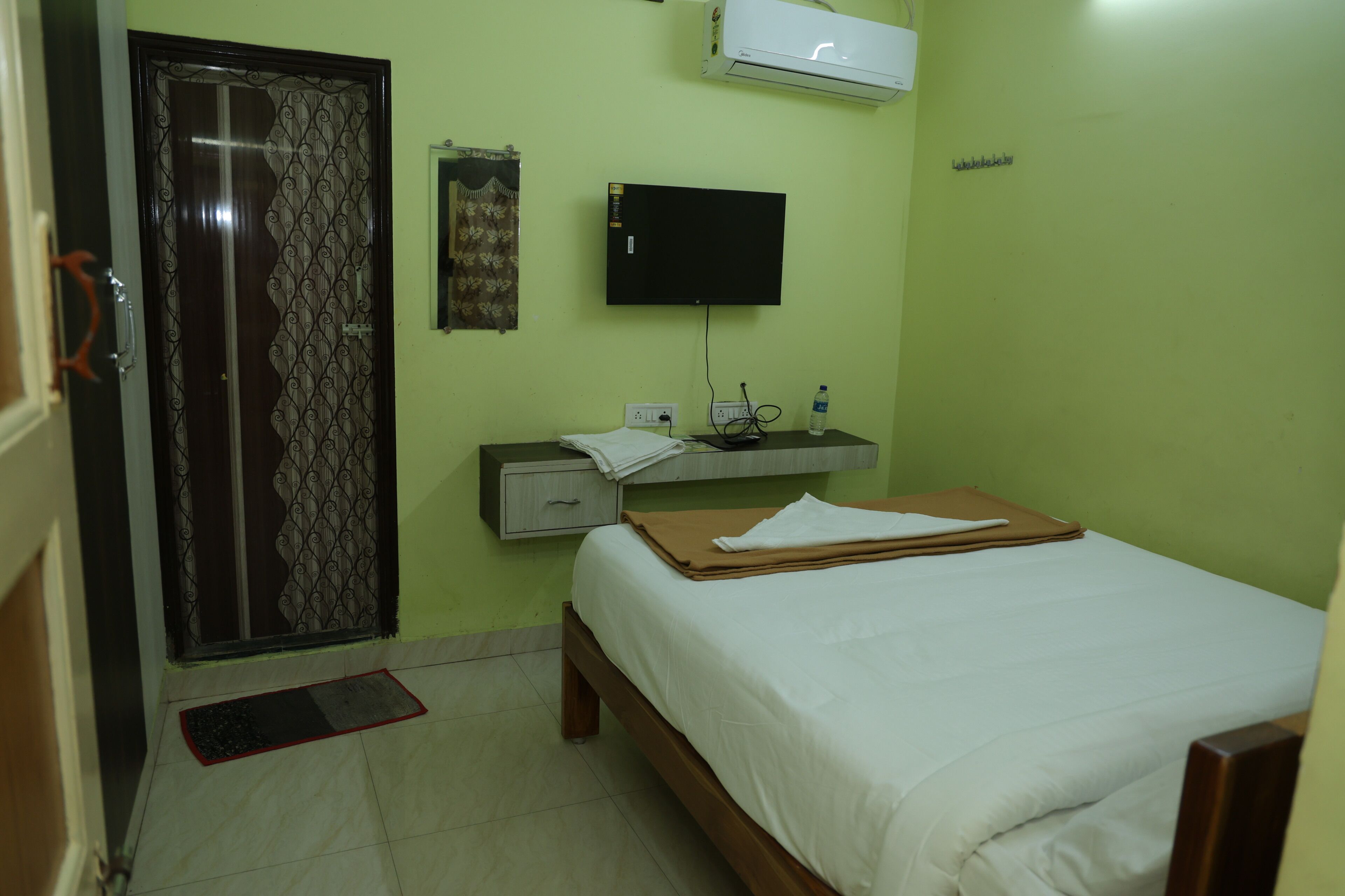 A room with a bed, a desk with a flat-screen TV, a wall-mounted air conditioner, a door with a decorative pattern, and a small bedside table with a water bottle.