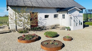 A white building with a metal roof, a wooden door, and a small window. There are garden beds with plants and a gravel driveway. - Rural house in a peaceful location with wonderful views of the Scanian plains. (Svedala)