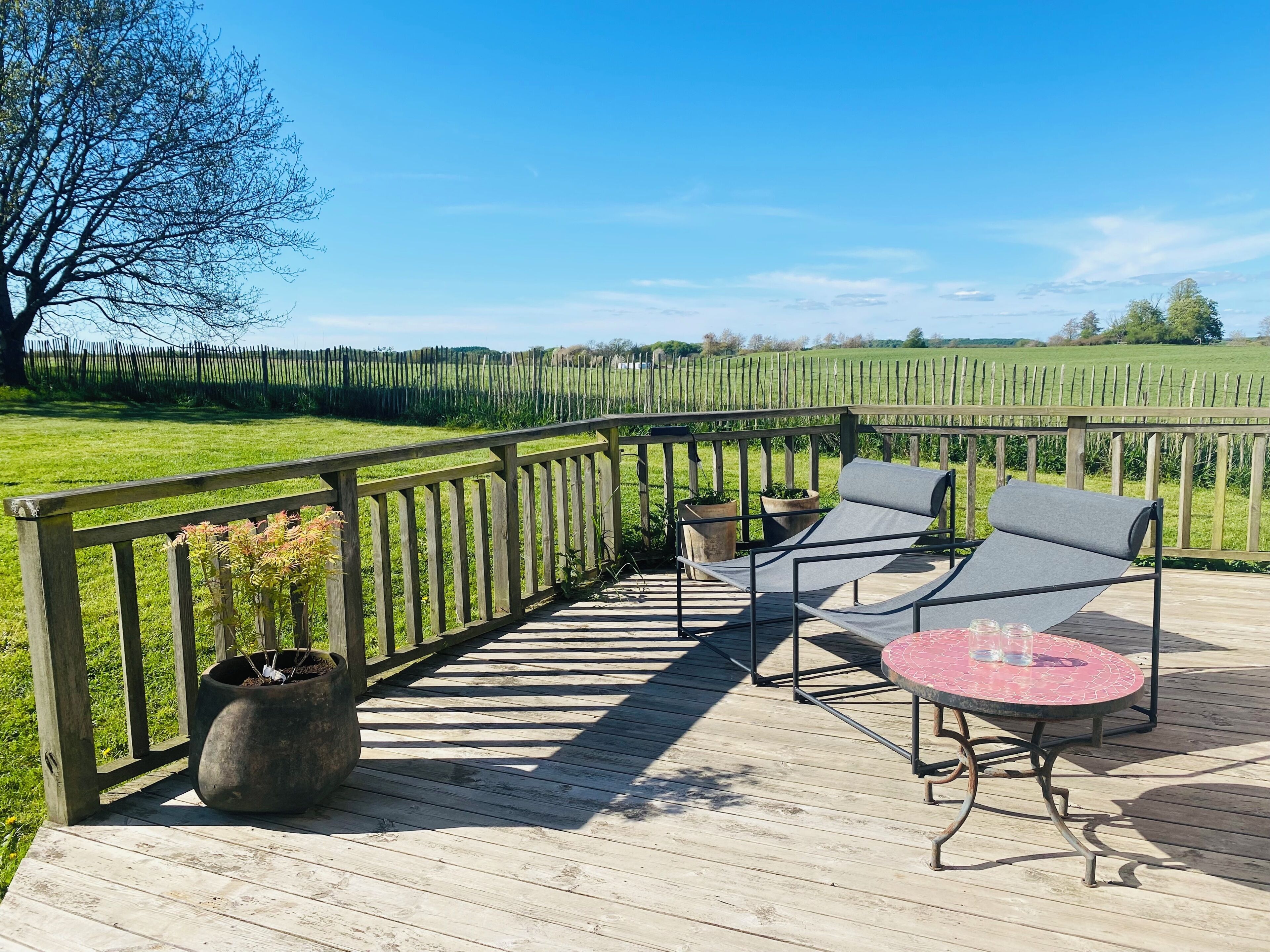 A wooden deck with a metal railing, two lounge chairs, a small table, and potted plants.