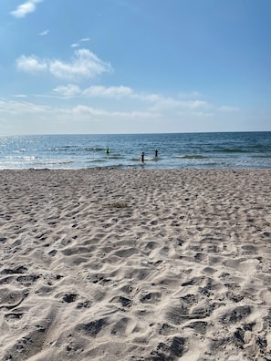 En sandstrand med fotspår som leder ner mot vattnet, en klar himmel och ett lugnt hav.