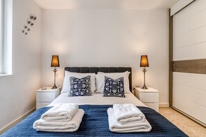 A neatly made bed with two pillows, two bedside lamps, and a stack of towels on a blue quilted bedspread. - Modern Surrey Quays Flat (London)