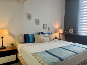 Ein Schlafzimmer mit einem Bett, zwei Nachttischlampen, einem Nachttisch mit Pflanze und an der Wand befestigten Bildern.