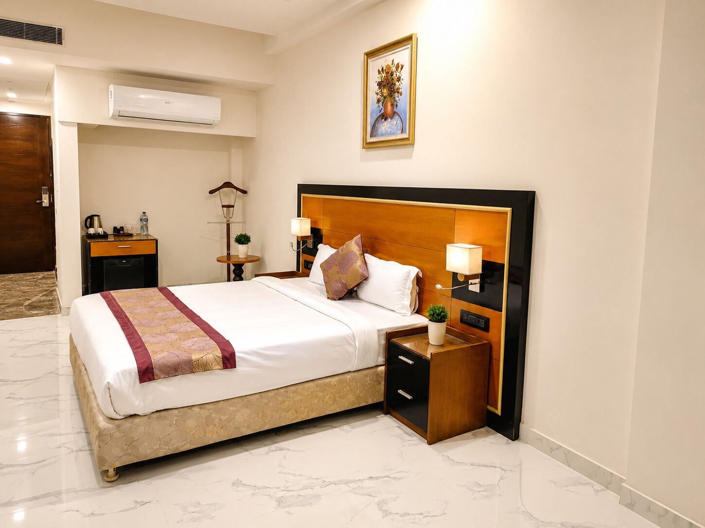 A hotel room with a bed, bedside tables, a nightstand, a lamp, a wall-mounted air conditioner, and a painting.