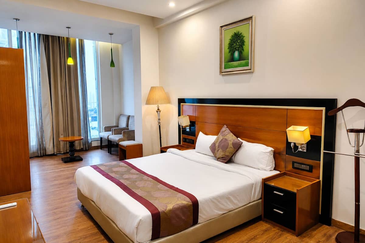 Quarto de hotel com uma cama grande, mesas de cabeceira, escrivaninha, uma cadeira, abajur e um quadro na parede.