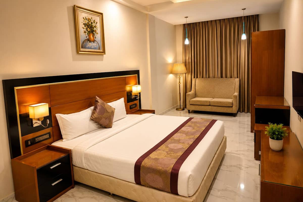 Quarto de hotel com cama, mesas de cabeceira, luminária, poltrona, televisão e planta.