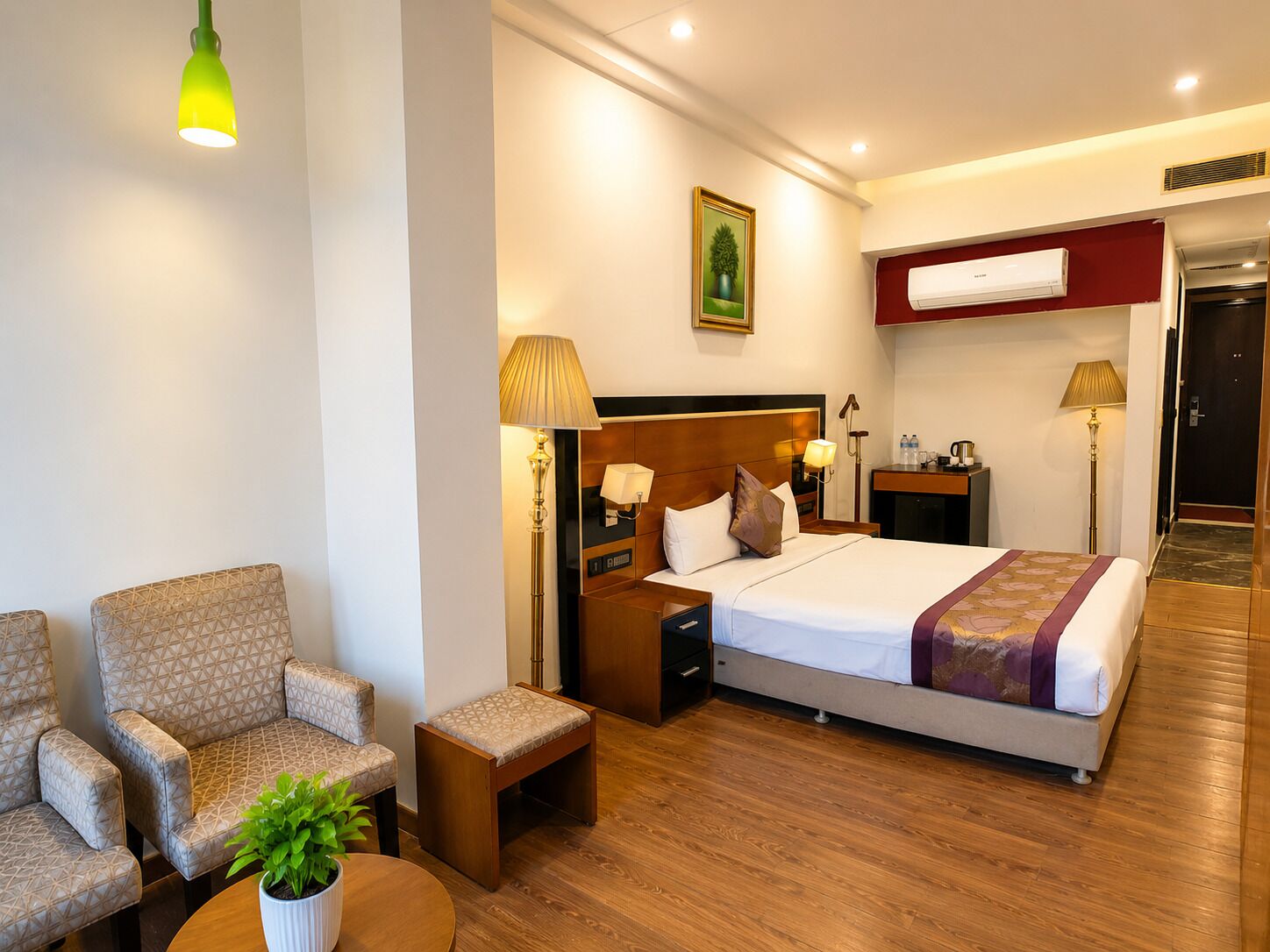 Una habitación de hotel con una cama, dos sillones, una mesita de noche, una lámpara, una pequeña mesa con una planta y un aire acondicionado instalado en la pared.
