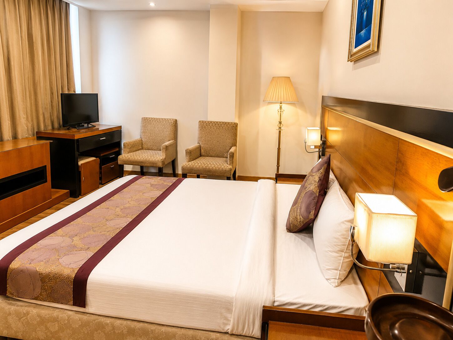 A hotel room with a bed, two armchairs, a desk with a television, a lamp, and a painting on the wall.