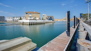 Una propiedad frente al agua con un muelle de madera que se adentra en una superficie acuática tranquila, rodeada de edificios modernos y un cielo despejado.