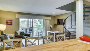 Un vaste espace de vie comprenant une table à manger en bois, des sièges confortables, un escalier et une grande fenêtre donnant sur l’extérieur.